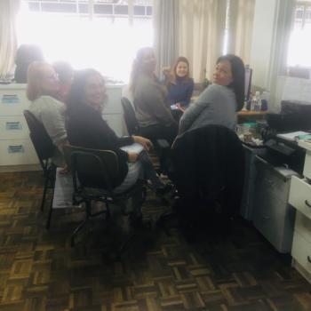 Imagem de seis mulheres sentadas em grupo, olhando para trs e sorrindo para a foto. Elas esto numa sala de escritrio, com mveis brancos no entorno delas.