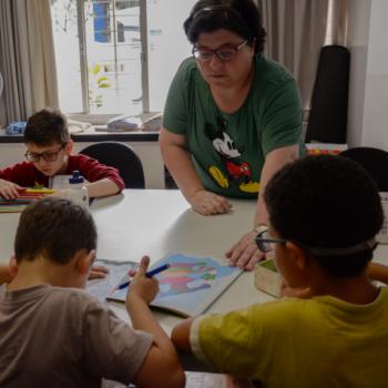 a imagem mostra trs alunos crianas e uma professora em volta de uma mesa branca. Um aluno est de frente para a foto, sentado, pegando lpis de cor numa caixa. Ele tem  sua esquerda (direita da foto) a professora, que est em p olhando e segurando a pontinha do caderno de desenhos que est sendo usado por um outro estudante, sentado  sua frente, do outro lado da mesa, de costas para a foto. Ele est de cabea baixa, desenhando. Ao lado direito dele, tambm de costas para a foto, h outro aluno, tambm sentado, que olha para o desenho do colega  esquerda dele.